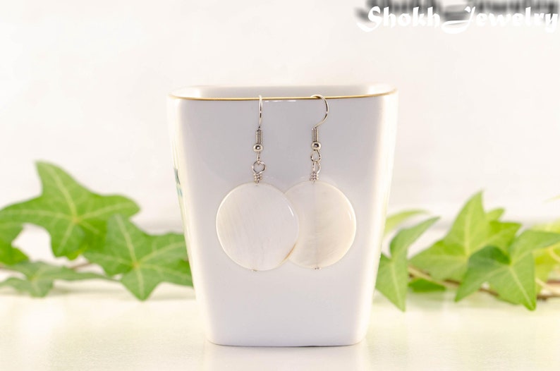 May include: A pair of white shell earrings with silver hooks. The earrings are round and have a natural, organic texture.