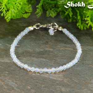 May include: A delicate bracelet made of small, round, opalescent beads. The bracelet is fastened with a silver clasp and a small charm. The background features green foliage and the text "Shokh Jewelry".