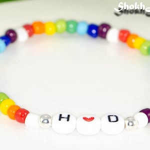 May include: A multi-coloured beaded bracelet with the letters "H❤️D" on white beads. The bracelet displays a spectrum of colours, including red, orange, yellow, green, blue, and purple. The bracelet is a piece of jewellery.