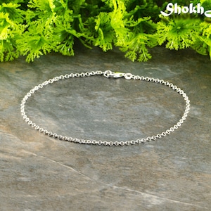 May include: A silver chain anklet with a lobster clasp fastening. The anklet is lying on a grey textured surface.