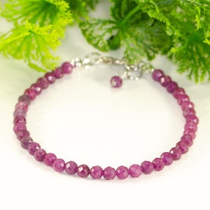 May include: A delicate bracelet featuring faceted, deep pink gemstones strung together with a silver-toned chain. The bracelet is displayed against a backdrop of green foliage and the text "Shokh Jewelry".