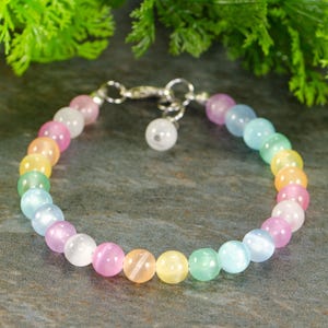 Peut inclure: Un bracelet de perles multicolores avec des perles pastel roses, blanches, jaunes, vertes et bleues. Le bracelet est doté d'un fermoir argenté et d'un petit charme blanc. La marque "Shokh Jewelry" est visible en arrière-plan.
