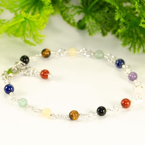 May include: A silver chain bracelet with a selection of colourful gemstone beads. The bracelet features round beads in colours such as red, blue, yellow, and black. The bracelet is displayed on a white surface with green foliage in the background. The text "Shokh Jewelry" is visible.