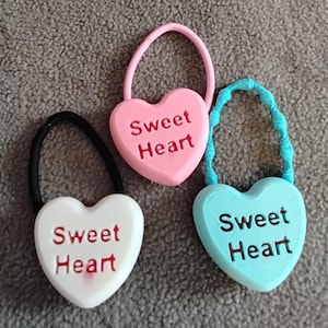 May include: Three heart-shaped hair ties in pink, white, and blue. Each heart has the words "Sweet Heart" in red. The hair ties have black, pink, and blue elastic bands.