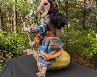 May include: A whimsical handmade witch doll riding a broom. The doll has a long nose, black hair, and is wearing a blue and orange patterned dress. The witch is perched on a yellow gourd, with a small sack on her back.