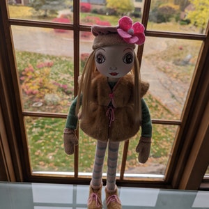 Puede incluir: Una mu&ntilde;eca con el pelo largo y rubio que lleva un chaleco de piel sint&eacute;tica marr&oacute;n, una diadema de flores rosa y leggings a rayas. La mu&ntilde;eca est&aacute; de pie frente a una ventana con vistas a un barrio suburbano.