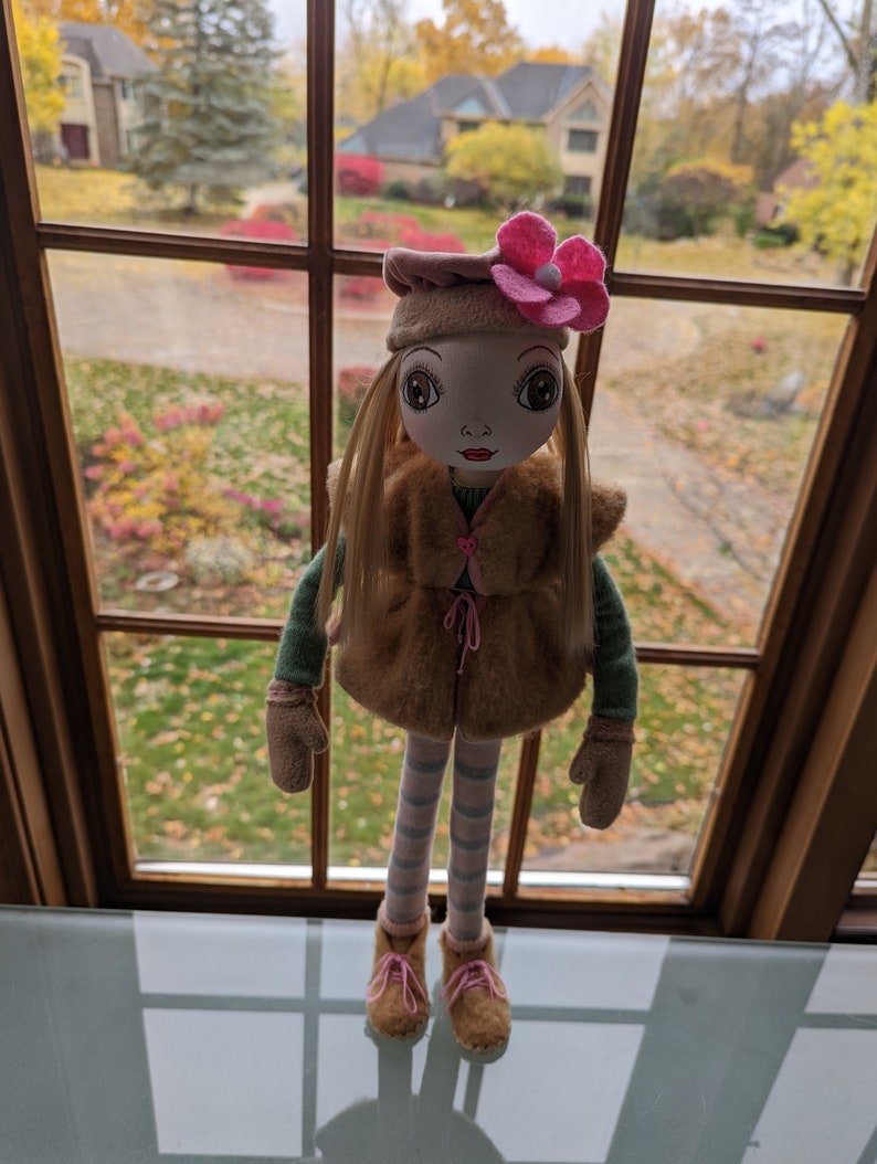 Puede incluir: Una mu&ntilde;eca con el pelo largo y rubio que lleva un chaleco de piel sint&eacute;tica marr&oacute;n, una diadema de flores rosa y leggings a rayas. La mu&ntilde;eca est&aacute; de pie frente a una ventana con vistas a un barrio suburbano.
