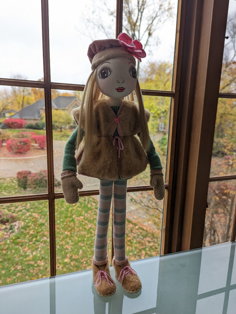 Puede incluir: Una mu&ntilde;eca de peluche con cabello rubio, que lleva un chaleco de piel marr&oacute;n, un su&eacute;ter a rayas verde y blanco, leggings a rayas rosa y blanco, botas marrones y una flor rosa en un sombrero marr&oacute;n.
