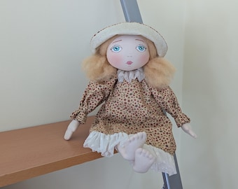 Könnte beinhalten: Eine handgemachte Puppe mit blonden Haaren, die einen weißen Hut, ein braunes und rotes Blumenkleid und einen weißen Spitzenbesatz trägt. Die Puppe hat blaue Augen und ein bemaltes Gesicht.