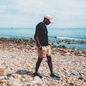 May include: A light pink baseball cap, a plain black t-shirt, and tan shorts featuring a colorful graphic print on the lower left leg. The shorts have a white drawstring at the waist. Dark blue slip-on sneakers are worn on the feet.