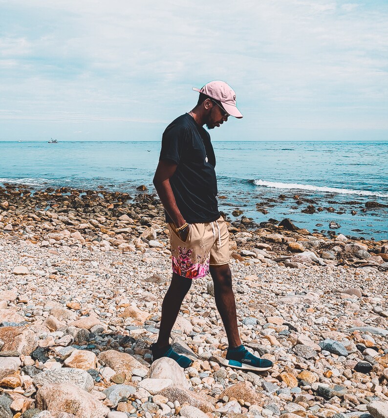 May include: A light pink baseball cap, a plain black t-shirt, and tan shorts featuring a colorful graphic print on the lower left leg. The shorts have a white drawstring at the waist. Dark blue slip-on sneakers are worn on the feet.