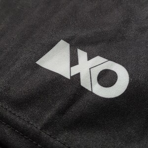 Op de afbeelding: Een zwart stoffen oppervlak met een wit grafisch logo. Het logo bestaat uit een effen driehoeksvorm links van de letters 'xo', waarbij de 'x' en de 'o' met elkaar verbonden zijn in een vetgedrukt, schreefloos typografisch ontwerp.