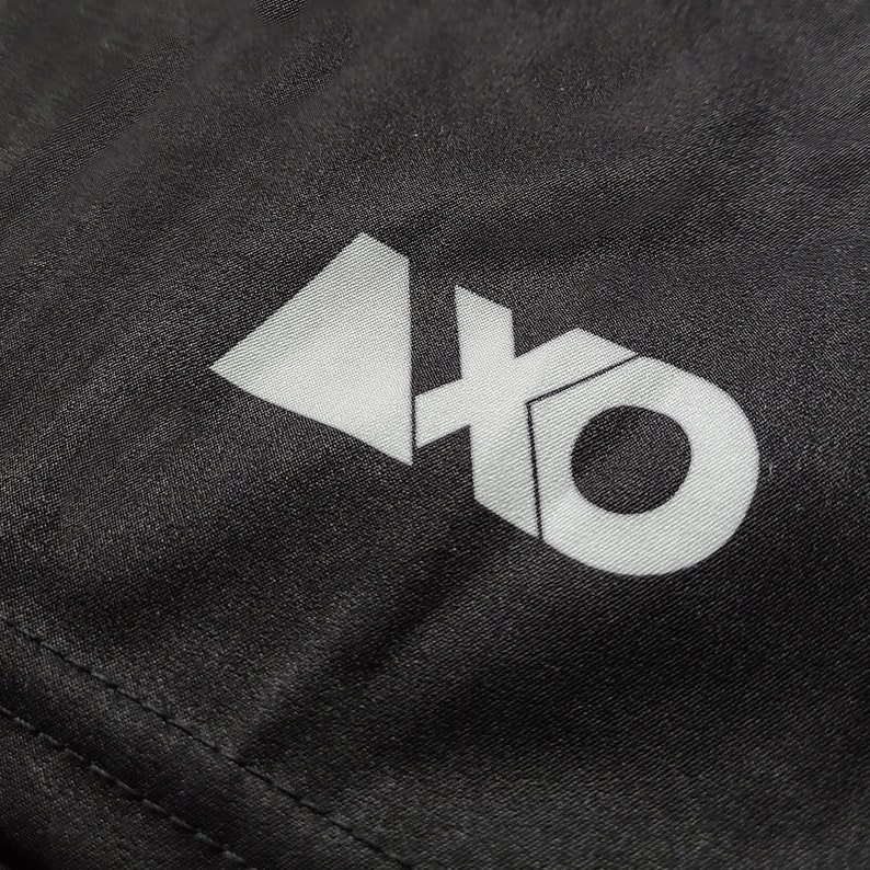 Op de afbeelding: Een zwart stoffen oppervlak met een wit grafisch logo. Het logo bestaat uit een effen driehoeksvorm links van de letters 'xo', waarbij de 'x' en de 'o' met elkaar verbonden zijn in een vetgedrukt, schreefloos typografisch ontwerp.