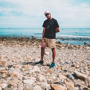 May include: A black t-shirt, tan shorts with a floral pattern on the left leg, a pink baseball cap, and dark sneakers. The shorts feature a small black logo on the right leg. The outfit is worn with dark sunglasses and a simple necklace.