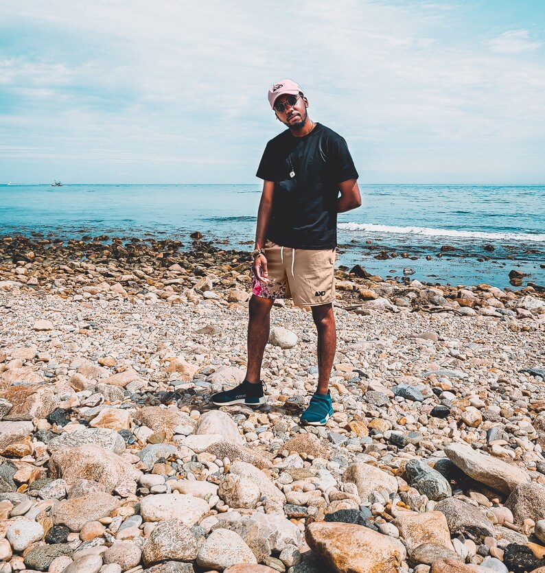 May include: A black t-shirt, tan shorts with a floral pattern on the left leg, a pink baseball cap, and dark sneakers. The shorts feature a small black logo on the right leg. The outfit is worn with dark sunglasses and a simple necklace.