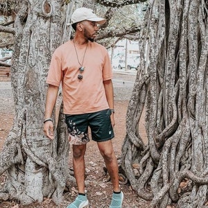 Op de afbeelding: Een beige honkbalpet, een perzikkleurig T-shirt met korte mouwen, een zwarte sportshort met een bloemenpatroon aan de linkerkant en lichtblauwe instap-sneakers. Een donkere kralenketting met een hanger hangt om de nek.