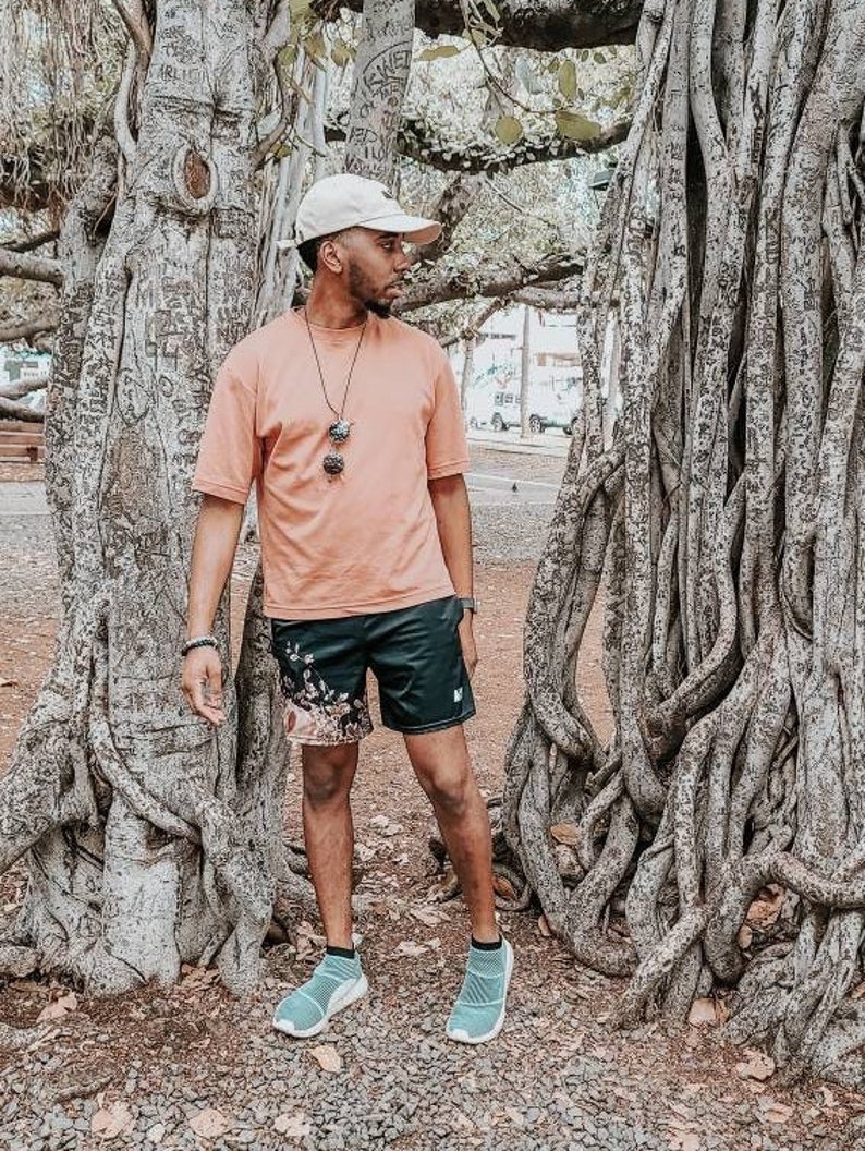Op de afbeelding: Een beige honkbalpet, een perzikkleurig T-shirt met korte mouwen, een zwarte sportshort met een bloemenpatroon aan de linkerkant en lichtblauwe instap-sneakers. Een donkere kralenketting met een hanger hangt om de nek.