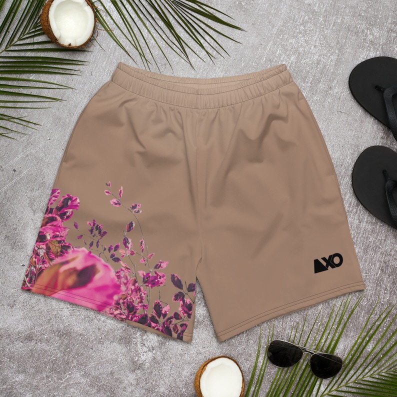 May include: Tan athletic shorts featuring an elastic waistband and a black logo on the right leg. The left leg displays a vibrant floral print with shades of bright pink, magenta, and dark purple leaves and petals against the solid tan fabric.