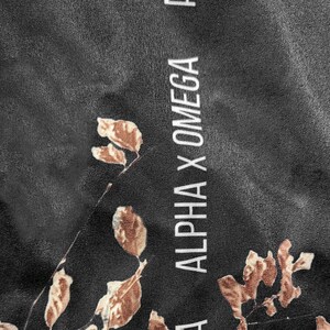 Op de afbeelding: Een zwarte stof met een herhalend patroon van bruine bladeren en de verticaal gedrukte witte tekst ALPHA X OMEGA. Het materiaal heeft een gladde, licht reflecterende textuur met een consistente donkere achtergrondkleur in het ontwerp.