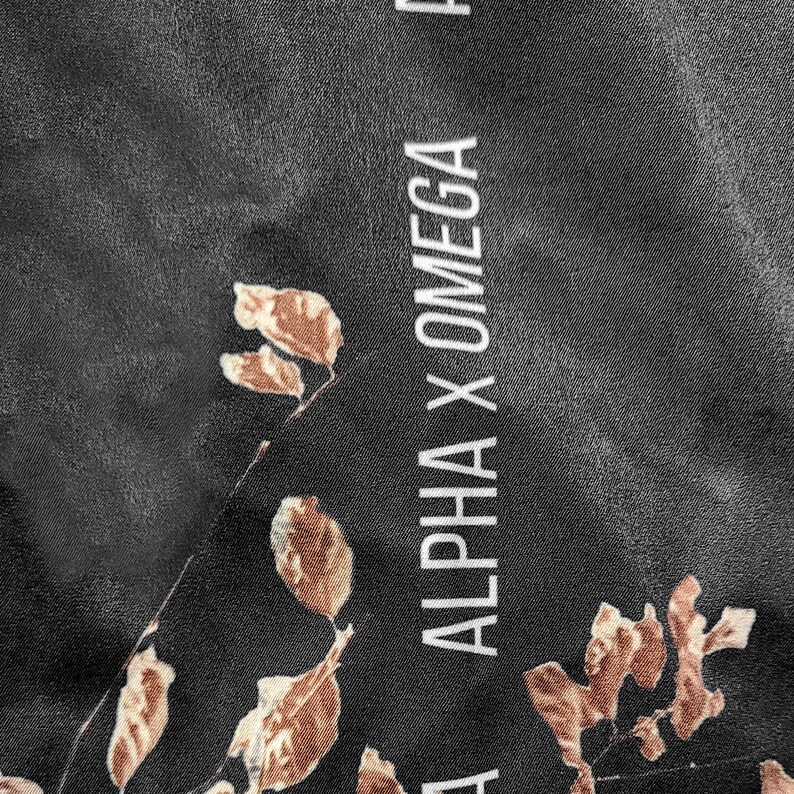 Op de afbeelding: Een zwarte stof met een herhalend patroon van bruine bladeren en de verticaal gedrukte witte tekst ALPHA X OMEGA. Het materiaal heeft een gladde, licht reflecterende textuur met een consistente donkere achtergrondkleur in het ontwerp.