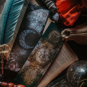 May include: A close-up of three vintage-style bookmarks with intricate designs. One bookmark features a crescent moon, another has a circular design with a compass, and the third has a floral pattern. The bookmarks are on a table with a book, a feather, and a skull.