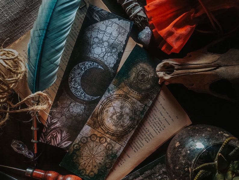 May include: A close-up of three vintage-style bookmarks with intricate designs. One bookmark features a crescent moon, another has a circular design with a compass, and the third has a floral pattern. The bookmarks are on a table with a book, a feather, and a skull.