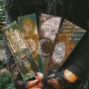 May include: Four vintage-style bookmarks with different designs. One features a hand with a palm reading, another has a moon and stars, a third has a compass and a fourth has a quote about the moon. The bookmarks are made of paper and have a distressed look.