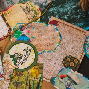 May include: Assortment of decorative paper ephemera with whimsical fairy and floral designs. The paper ephemera is in shades of green, blue, brown, and white.