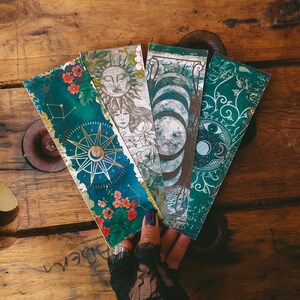 K&ouml;nnte beinhalten: Vier dekorative Lesezeichen mit himmlischen und floralen Designs. Die Lesezeichen sind in Blaut&ouml;nen, Gr&uuml;n, Gold und Rot gehalten. Die Lesezeichen werden in einer Hand gehalten, die einen schwarzen Spitzenhandschuh tr&auml;gt.