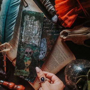 May include: A vintage style bookmark featuring a hand with lines and symbols, a skull with flowers, and a moon and stars design. The bookmark is on a book with a feather and other objects.