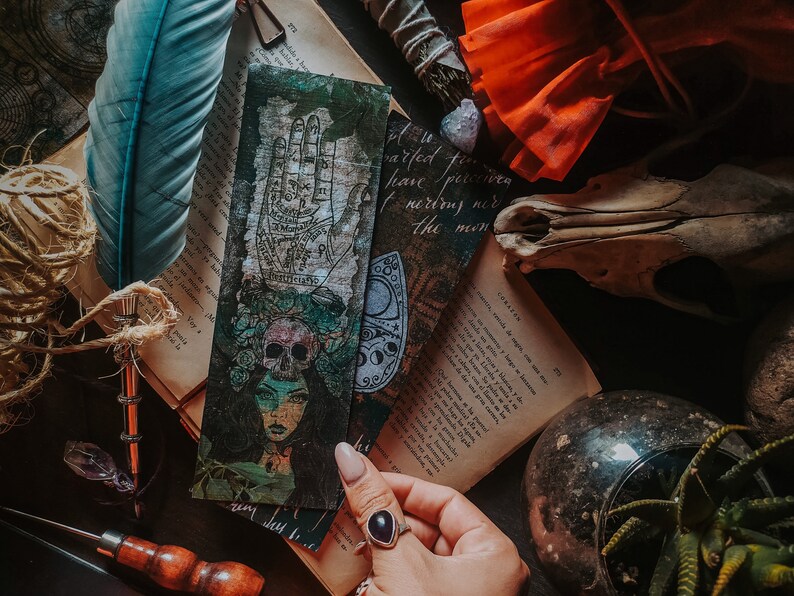 May include: A vintage style bookmark featuring a hand with lines and symbols, a skull with flowers, and a moon and stars design. The bookmark is on a book with a feather and other objects.