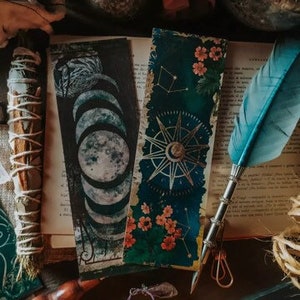 K&ouml;nnte beinhalten: Zwei dekorative Lesezeichen mit Vintage-&Auml;sthetik. Ein Lesezeichen zeigt ein Mondphasen-Design mit schwarzem Hintergrund und silbernen Akzenten. Das andere Lesezeichen hat einen marineblauen Hintergrund mit goldenen Akzenten und ein Kompass-Design mit einem floralen Muster.