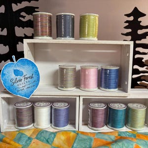 May include: Twelve spools of sewing thread in various colors including pink, blue, green, white, and grey, arranged on white wooden shelves. A blue leaf-shaped sticker with the text Silver Forest Quilts is attached to the side of the shelves.