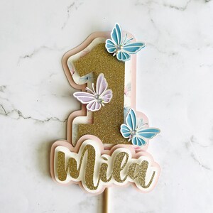 May include: A layered cake topper with the number "1" in gold glitter, adorned with light blue and lavender butterflies. The name "Mila" is written in gold glitter script below the number. The topper is on a wooden stick.