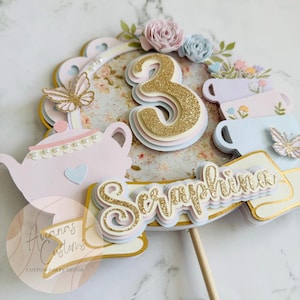 May include: A layered cake topper with a gold glitter number 3, the name "Seraphina", and a teapot design. The topper includes pastel colors, paper flowers, and butterfly accents. The design is for a party.