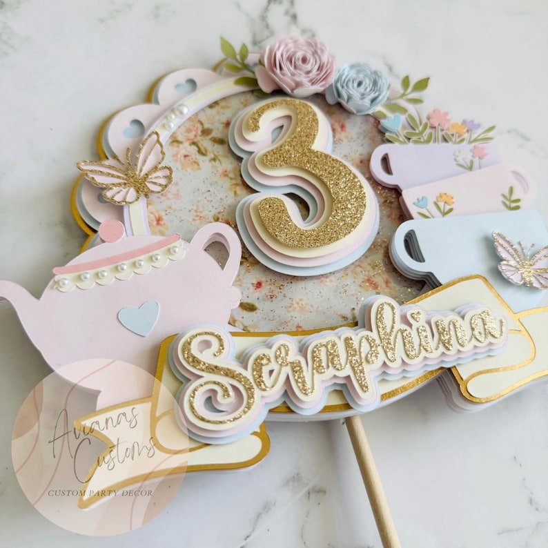 May include: A layered cake topper with a gold glitter number 3, the name "Seraphina", and a teapot design. The topper includes pastel colors, paper flowers, and butterfly accents. The design is for a party.