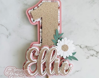 May include: A layered birthday cake topper with a large gold glitter number one. The number one is surrounded by a pink border and floral patterned paper. The name "Ellie" is also included in a layered design with gold glitter accents, along with a white daisy and green leaves.