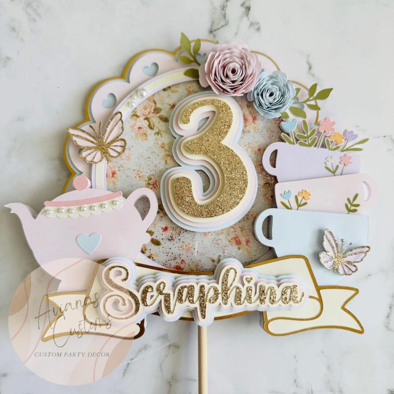 May include: A custom party decor cake topper with a floral theme. The topper features a gold glitter number 3, a pink teapot, teacups, butterflies, and the name "Seraphina" in gold glitter. The background has a floral pattern and the topper is on a wooden stick.