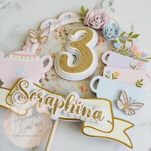 May include: A layered cake topper with a gold glitter number 3, floral accents, and a banner that reads "Seraphina." The design includes a teapot, teacups, and butterflies in pastel colors, perfect for a third birthday celebration.
