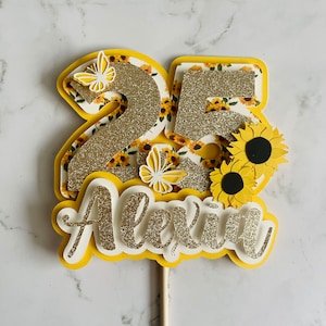 May include: A layered cake topper with the number "25" in gold glitter, sunflower and butterfly accents, and the name "Alexin". The topper is yellow, white, and gold, with a wooden stick for insertion into a cake.