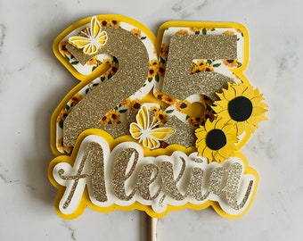May include: A layered cake topper with the number "25" in gold glitter, sunflower and butterfly accents, and the name "Alexin". The topper is yellow, white, and gold, with a wooden stick for insertion into a cake.