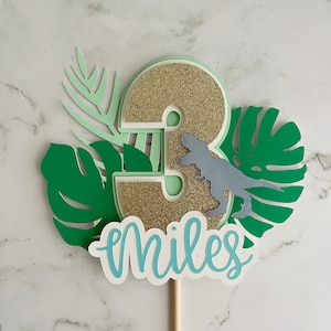 May include: A layered cake topper featuring a gold glitter number 3, a gray dinosaur silhouette, and green and light green paper leaves. The name "Miles" is written in a cursive font with a blue outline. The topper is attached to a wooden stick.