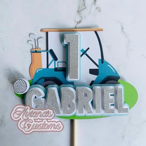 May include: A custom cake topper featuring a golf cart, golf ball, and the name "Gabriel" in silver glitter. The number "1" is also in silver glitter, indicating a first birthday. The topper includes the text "Aviana's Customs".