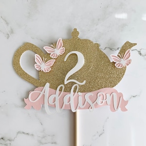May include: A gold glitter teapot-shaped cake topper with the number 2 and the name "Addison" in white script. Pink butterflies and a pink banner add a delicate touch to this birthday decoration.