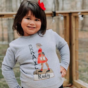 May include: A gray sweatshirt with long sleeves featuring the text "Brave like Ruby" and an illustration of a girl in a red dress holding books. The girl is standing on a stack of books. A red bow is in the person's hair.