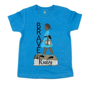 May include: A blue t-shirt featuring the words "BRAVE" and "like Ruby" with an illustration of a girl in a blue dress holding a book. The girl is standing on a stack of books. The shirt has short sleeves.