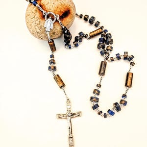 May include: A rosary featuring cylindrical and round beads in shades of metallic bronze and deep blue, connected by silver-toned metal links. The piece includes a silver-toned crucifix and a central medal depicting a figure in a circular frame.