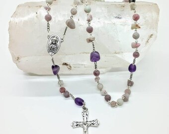 May include: A silver rosary with a silver crucifix and a silver charm featuring a religious figure. The beads are made of amethyst and pink and white stones.