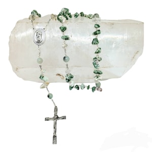 May include: A silver rosary with green and white stone beads. The rosary is displayed on a clear quartz crystal.