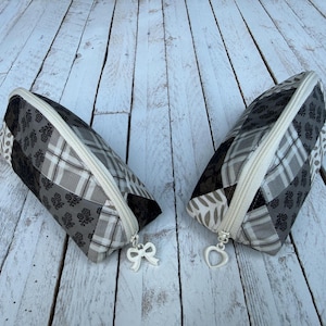 May include: Two zippered pouches featuring a patchwork design in shades of black, gray, and white. The fabric patterns include floral motifs, plaid, and leaf prints. Each pouch has a white zipper closure with a white charm, one shaped like a bow and the other like a heart.